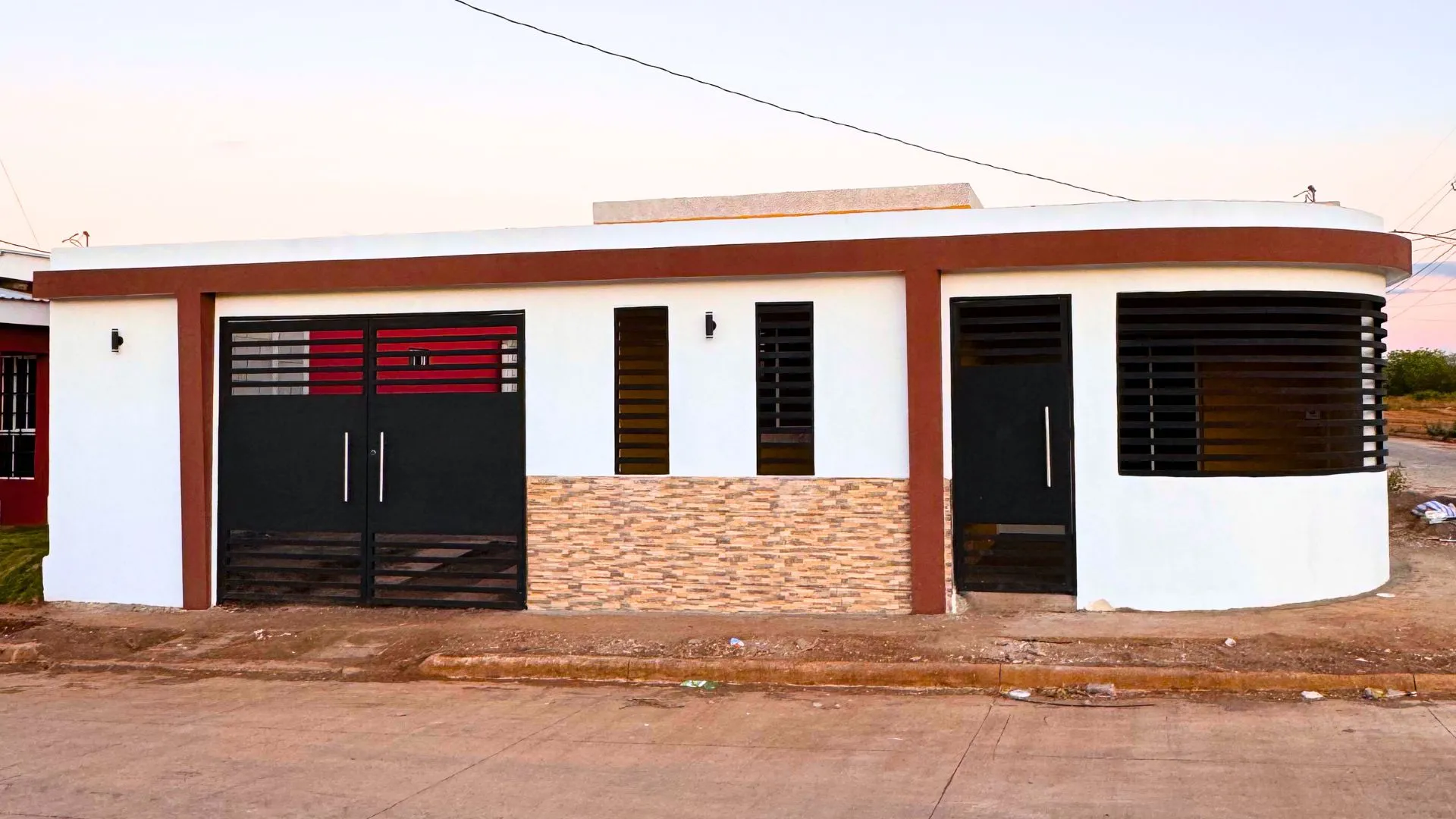 Muro frontal moderno y elegante construido con el sistema prefabricado Maxiblock. Presenta acabados en color blanco, molduras marrón oscuro, detalles de piedra decorativa texturizada, portón negro y una esquina curva con rejillas de ventilación integradas.