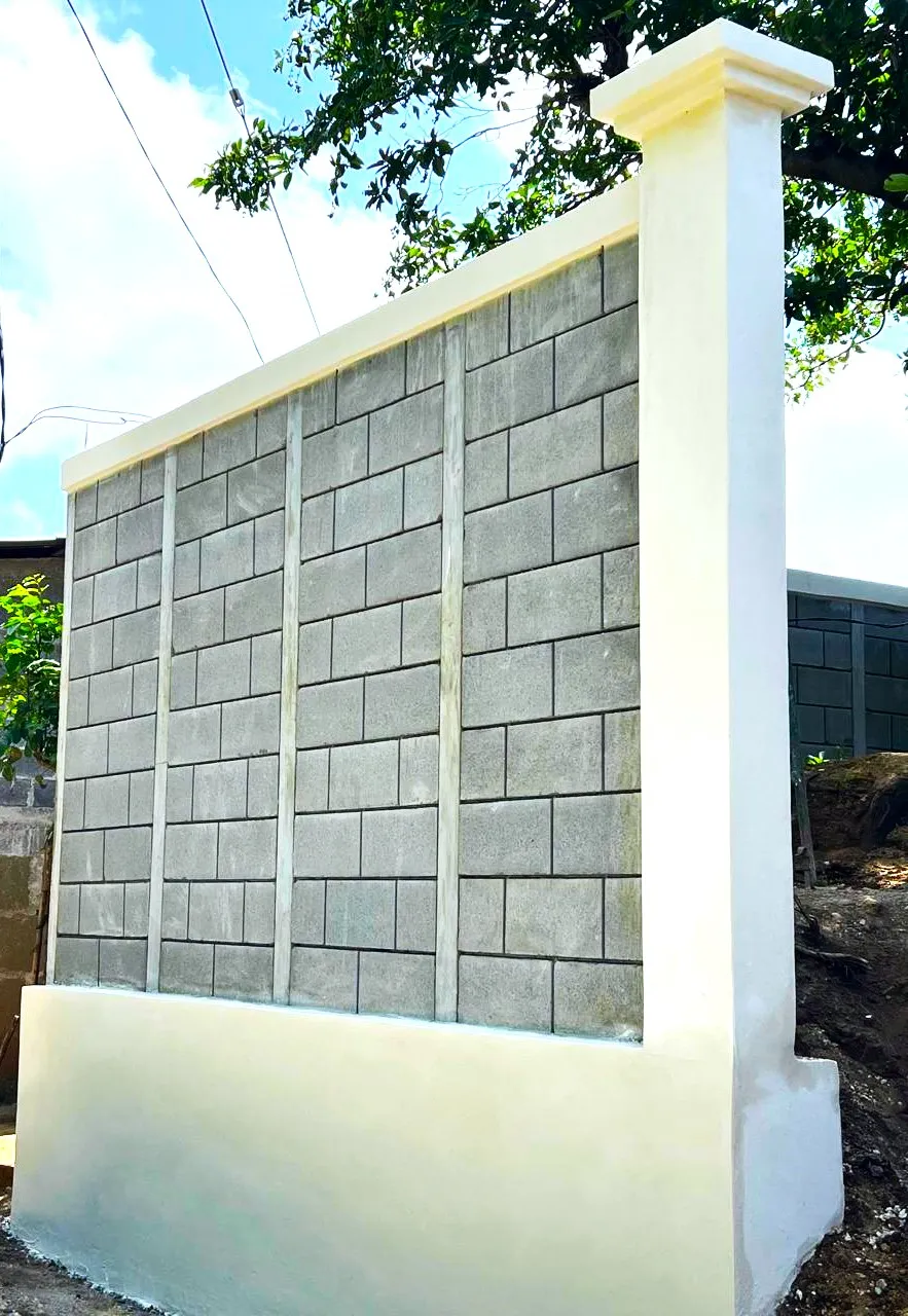 Sección de muro frontal compuesto construido con el sistema prefabricado Maxiblock. Muestra un zócalo en la base que funciona como muro de contención para retener el cambio de altura entre la calle y la terraza del terreno, complementado con una columna estilizada de concreto armado diseñada para sostener el portón.