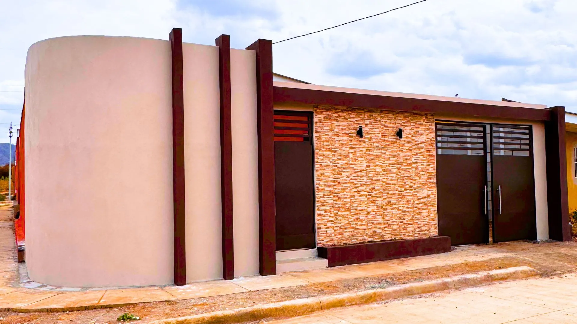 Fachada frontal residencial de estilo contemporáneo hecha con muros prefabricados Maxiblock. Destaca un revestimiento completo de piedra en tonos cálidos, portones metálicos en marrón chocolate y un diseño arquitectónico con tres columnas verticales decorativas en el extremo izquierdo.