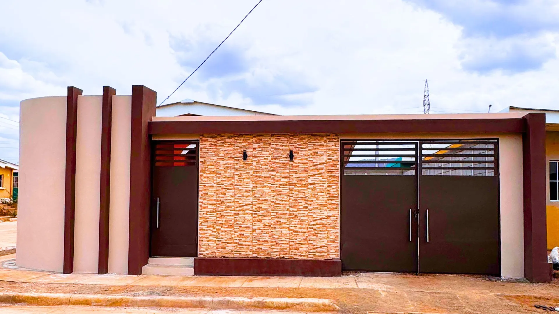 Fachada frontal residencial de estilo contemporáneo hecha con muros prefabricados Maxiblock. Destaca un revestimiento completo de piedra en tonos cálidos, portones metálicos en marrón chocolate y un diseño arquitectónico con tres columnas verticales decorativas en el extremo izquierdo.
