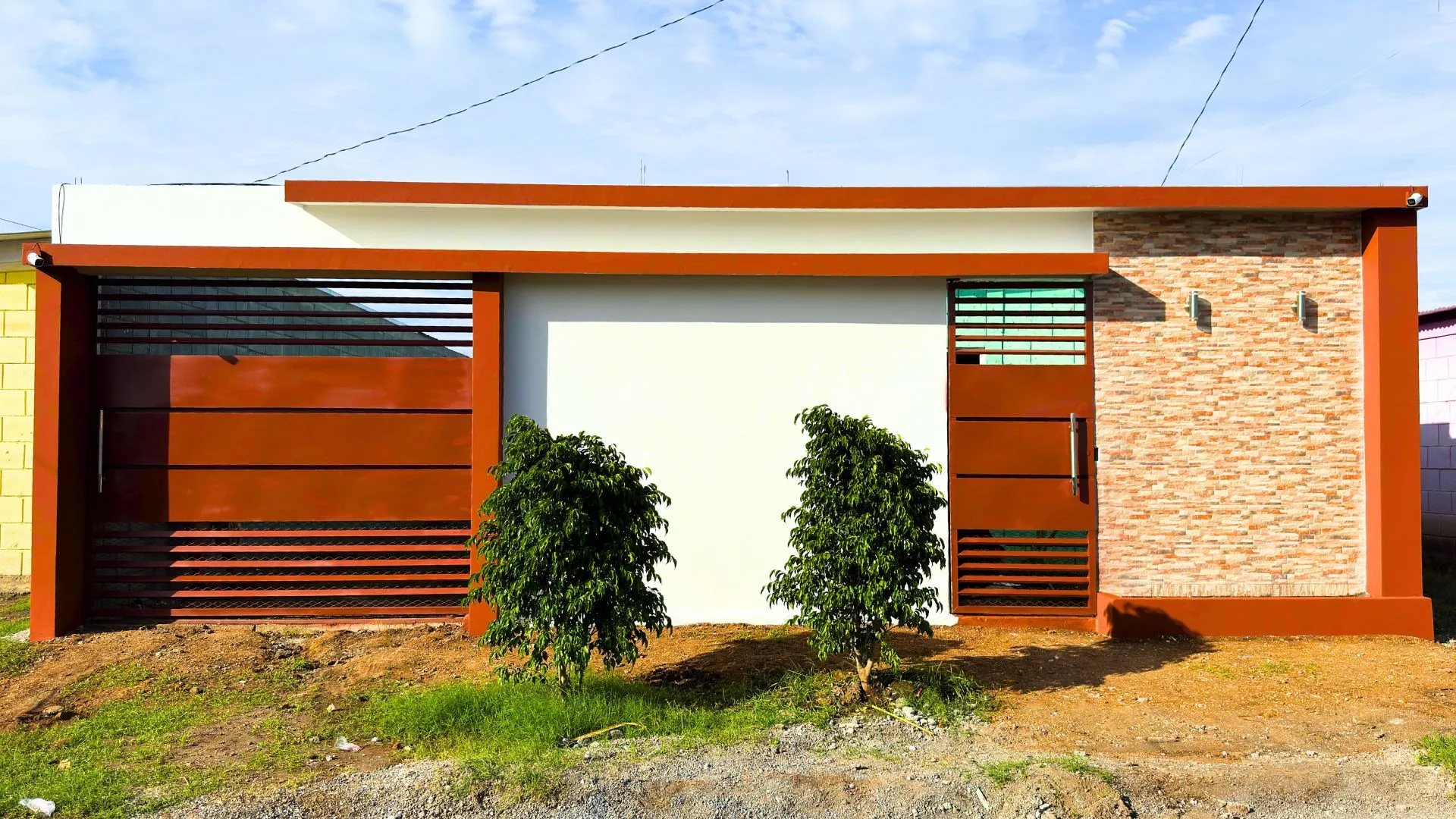 Diseño de muro perimetral frontal minimalista utilizando la tecnología constructiva Maxiblock. Muestra una combinación limpia de paredes blancas lisas, enchape de piedra rústica, vigas superiores en tono terracota y portones modernos de color óxido con líneas horizontales.