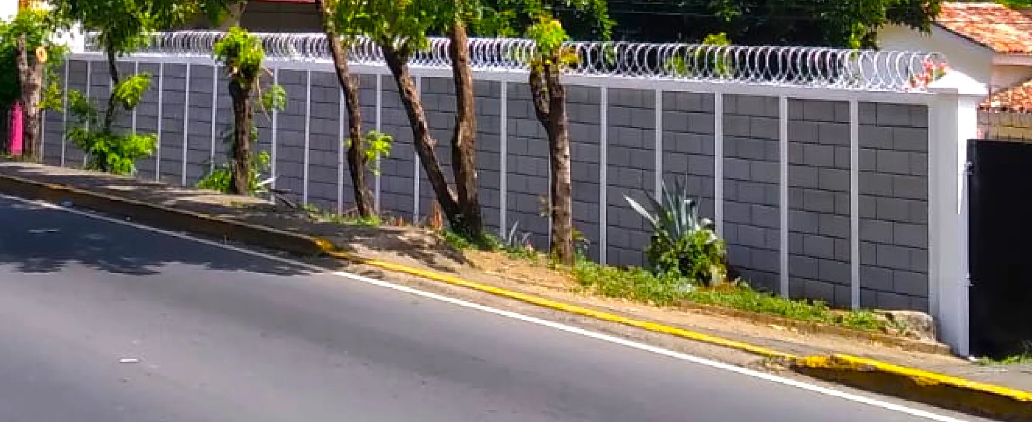 Extenso muro perimetral de seguridad para proyectos industriales o residenciales hecho con Maxiblock. Cuenta con columnas de concreto visto espaciadas uniformemente y protección superior de alambre de púas tipo concertina junto a una carretera.