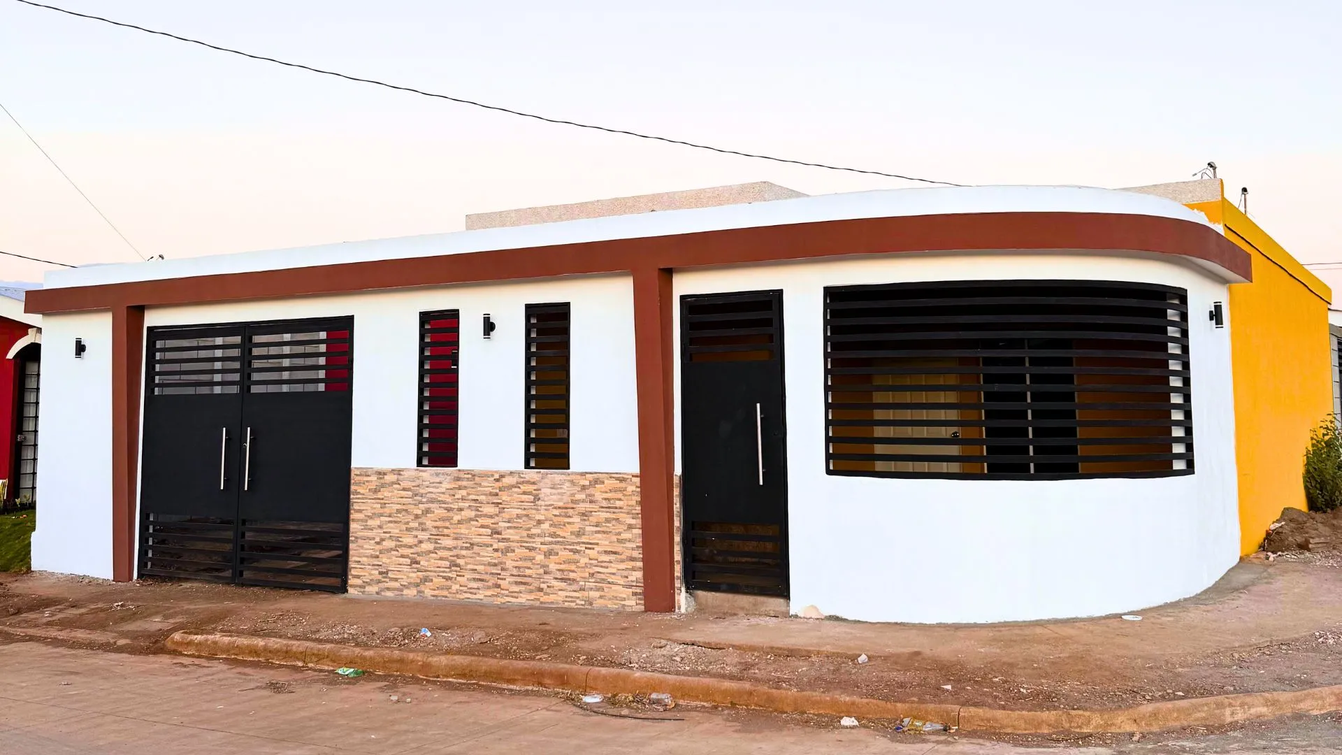 Muro frontal moderno y elegante construido con el sistema prefabricado Maxiblock. Presenta acabados en color blanco, molduras marrón oscuro, detalles de piedra decorativa texturizada, portón negro y una esquina curva con rejillas de ventilación integradas.