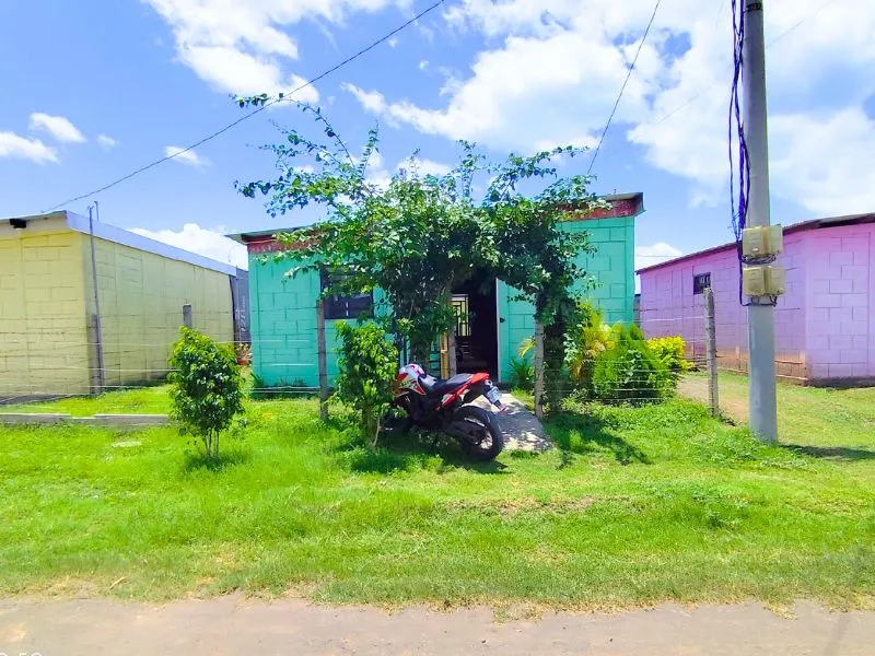 Fachada original antes de la renovación en Villa Flor de Pino, Managua. Se observa una vivienda sencilla con paredes de bloques prefabricados de color verde, un jardín frontal abierto con césped, árboles pequeños y una motocicleta estacionada en la entrada, delimitada por postes de concreto y alambre.