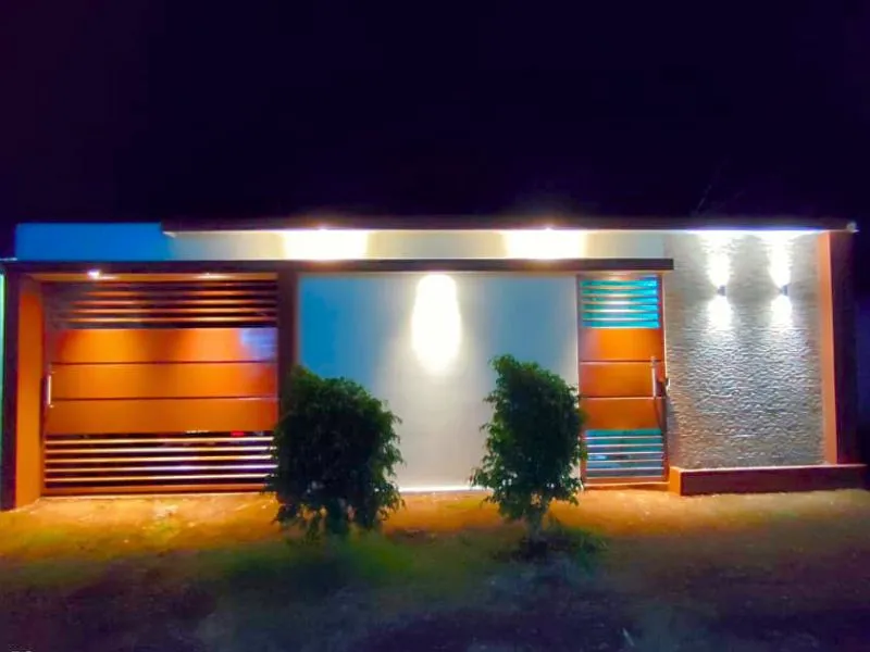 Fachada elegante terminada con iluminación nocturna en Villa Flor de Pino. La estructura terminada y pintada luce un portón y una puerta de estilo moderno iluminados por tiras LED superiores, una pared central con un reflector direccional y el muro de piedra texturizada resaltado por dos apliques de luz bidireccionales.