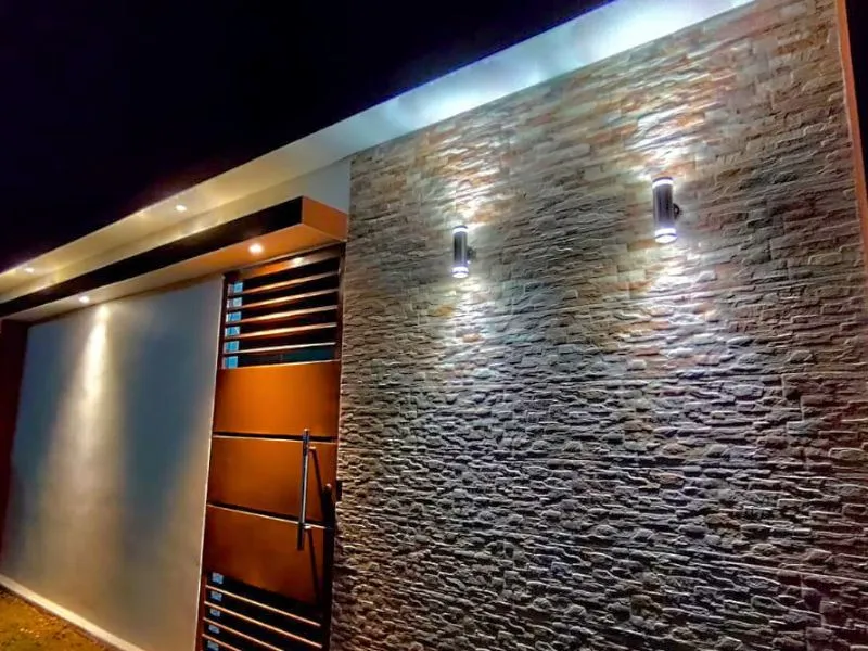 Primer plano nocturno del muro texturizado y los accesos de la fachada finalizada. Se aprecia a detalle el relieve del revestimiento de piedra iluminado por lámparas cilíndricas modernas, junto a la puerta peatonal metálica color marrón y los acabados de pintura blanca del portón principal.